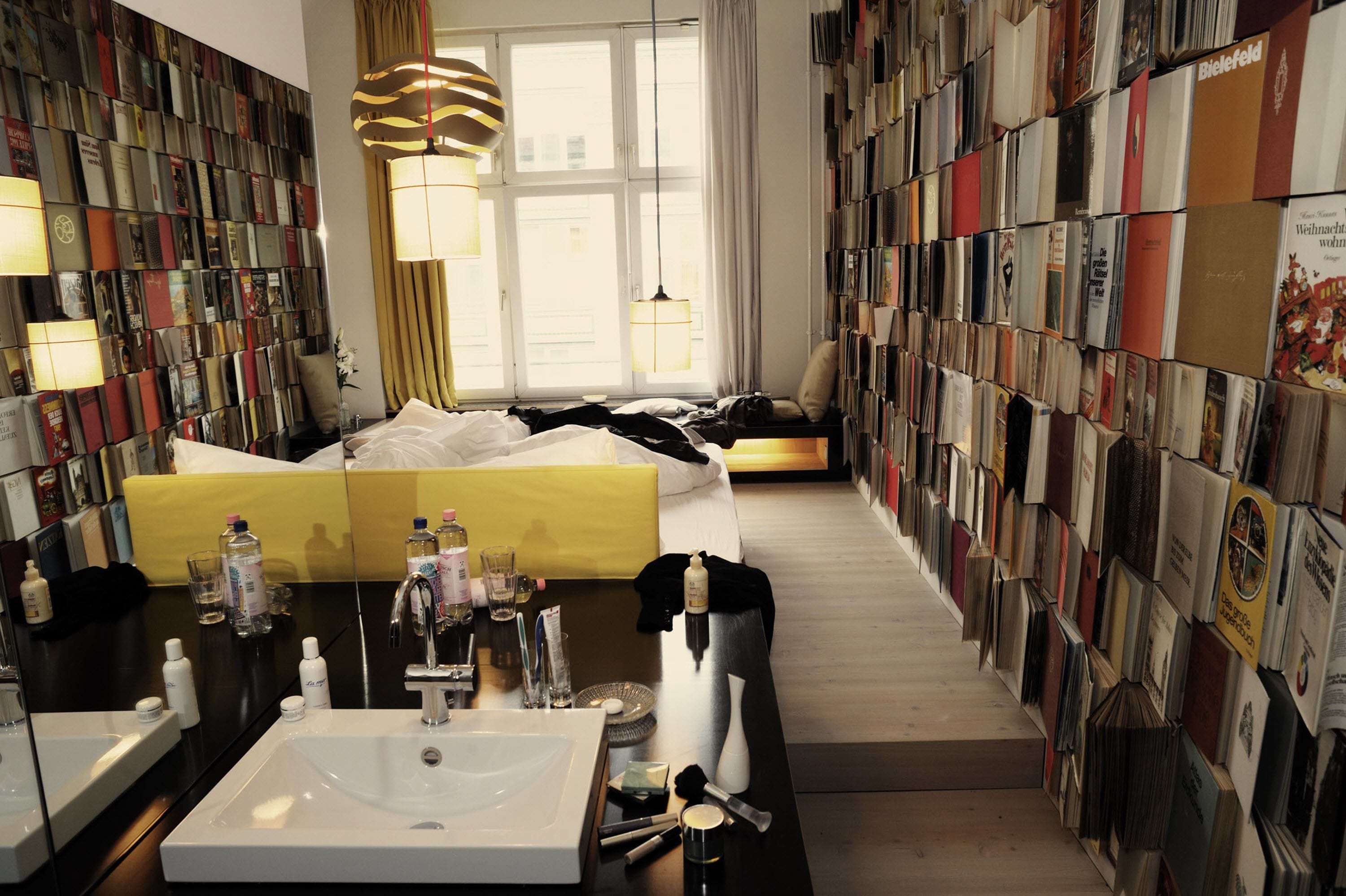 A hotel room with a large bed, a bedside table with toiletries, a wall of books, and a large window with curtains.