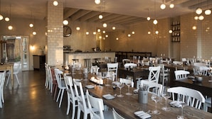 Un comedor amplio con varias mesas y sillas, un muro de ladrillo y lámparas colgantes.