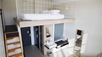 Una cama litera con colchón blanco, escalera de madera, un lavamanos y un espejo.