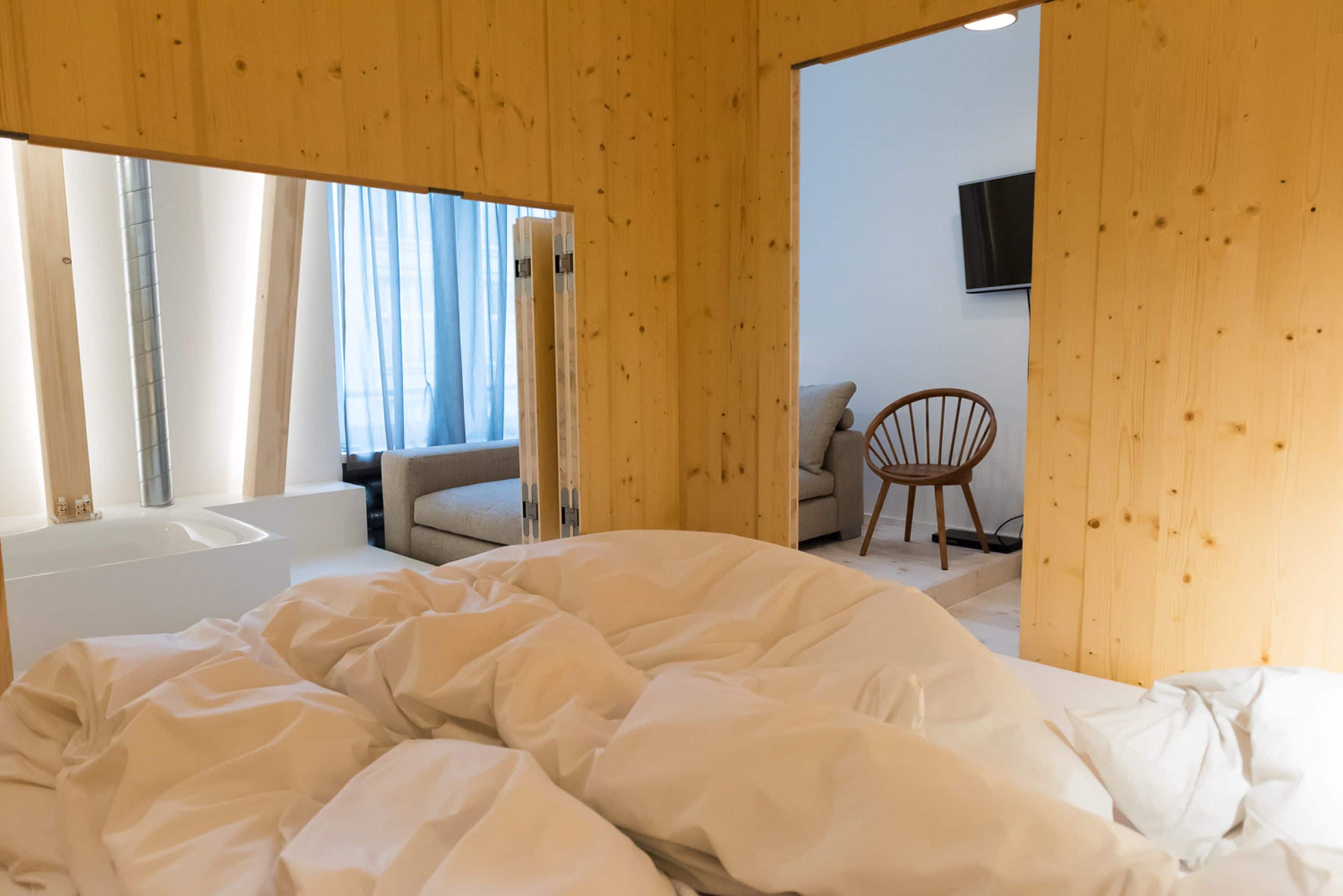 A bedroom with a large bed, a wooden ceiling, a window with curtains, a sofa, and a TV mounted on the wall.