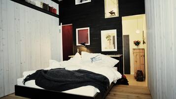 Un dormitorio con cama, piso de madera, pared blanca, puerta, espejo y estante con libros.