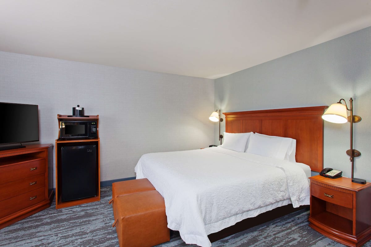 A hotel room with a bed, a television, a microwave, a refrigerator, a bedside table with a lamp, and a wooden nightstand.