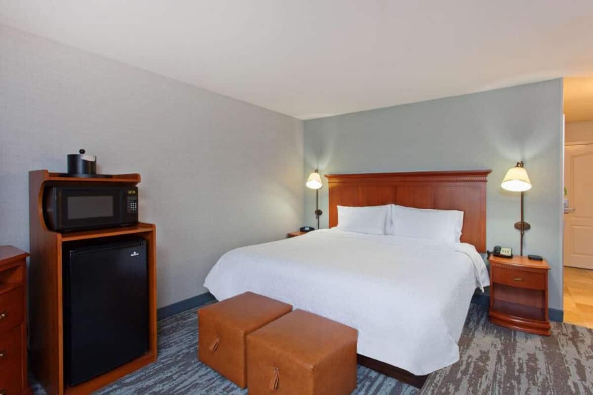 A hotel room with a bed, bedside lamps, a microwave, a refrigerator, and a small wooden bench.