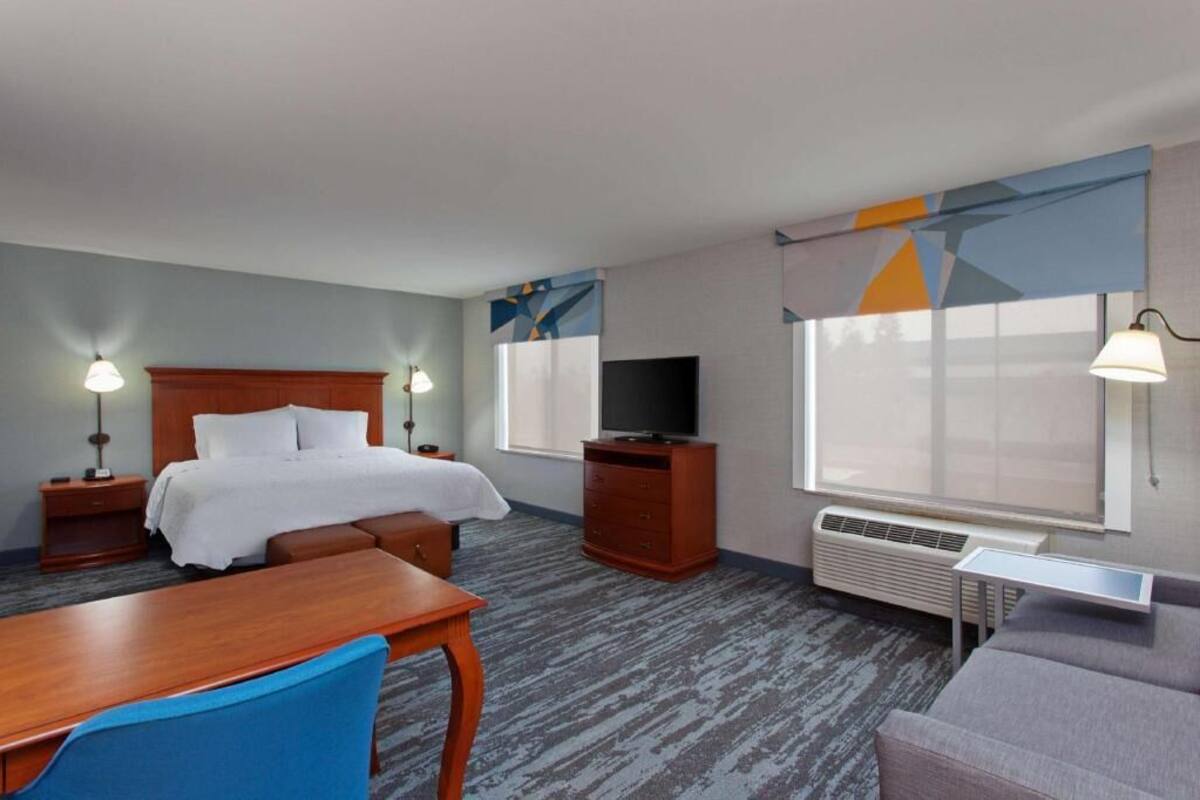A hotel room with a bed, a TV, a desk, a chair, a sofa, and a window with curtains.