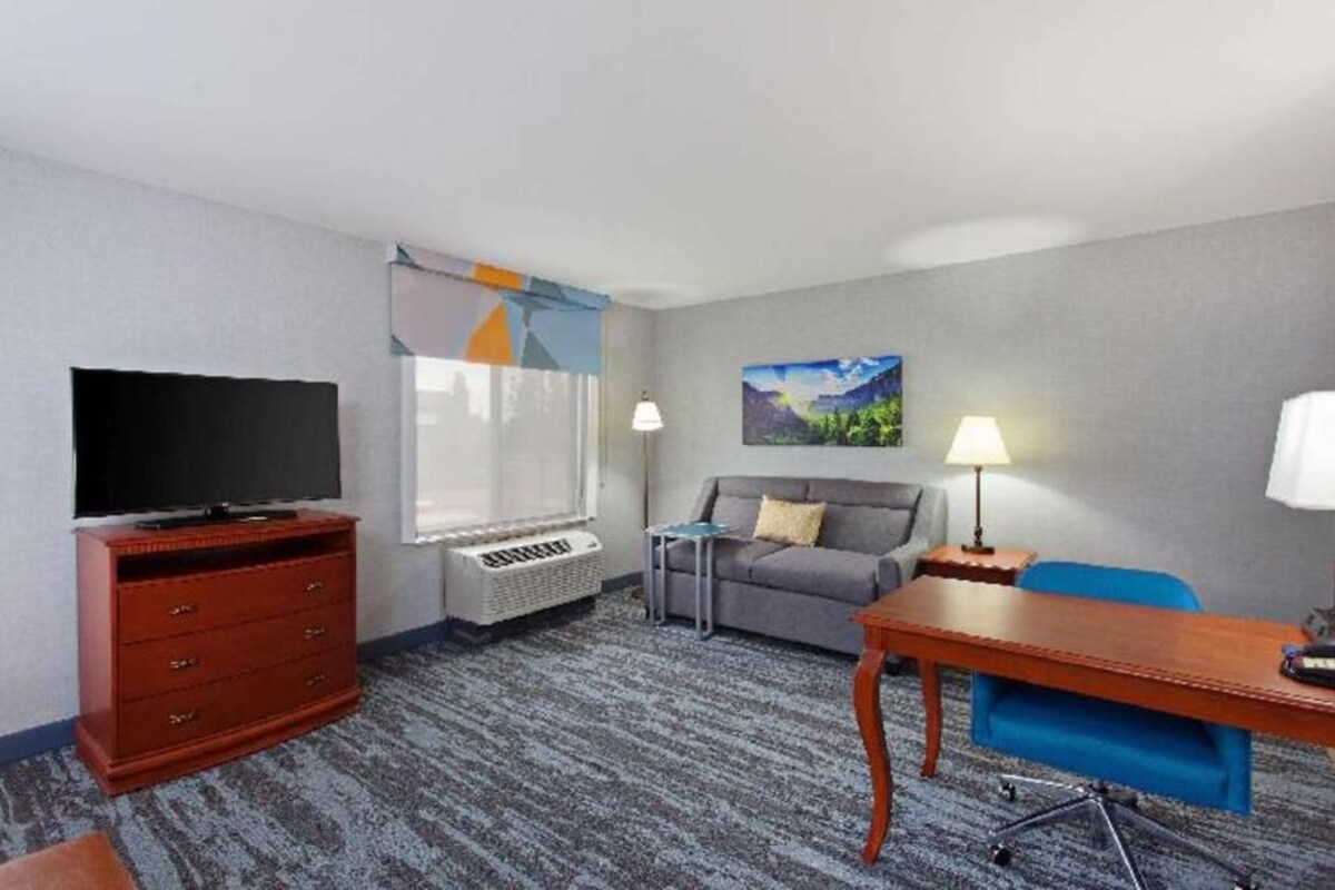 Habitación de hotel con televisión, sofá, escritorio, silla, lámpara y un cuadro en la pared.