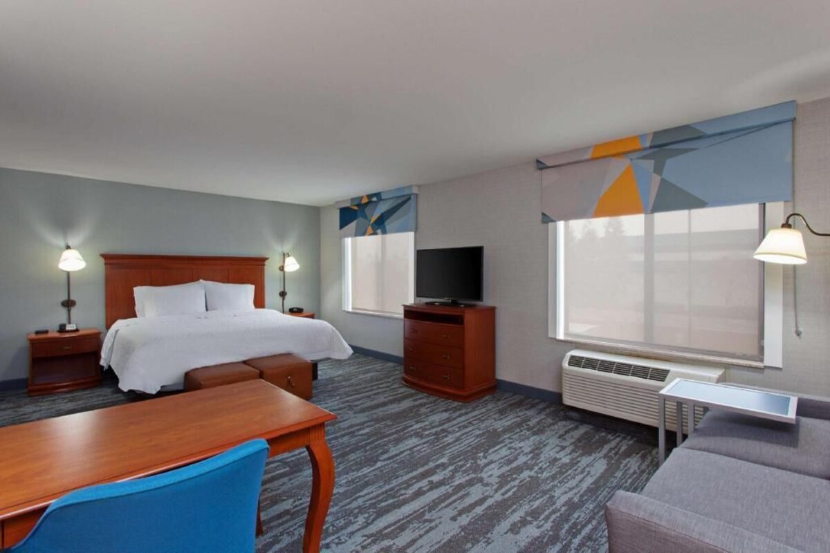 A hotel room with a bed, a TV, a desk, a chair, a sofa, and a window with curtains.