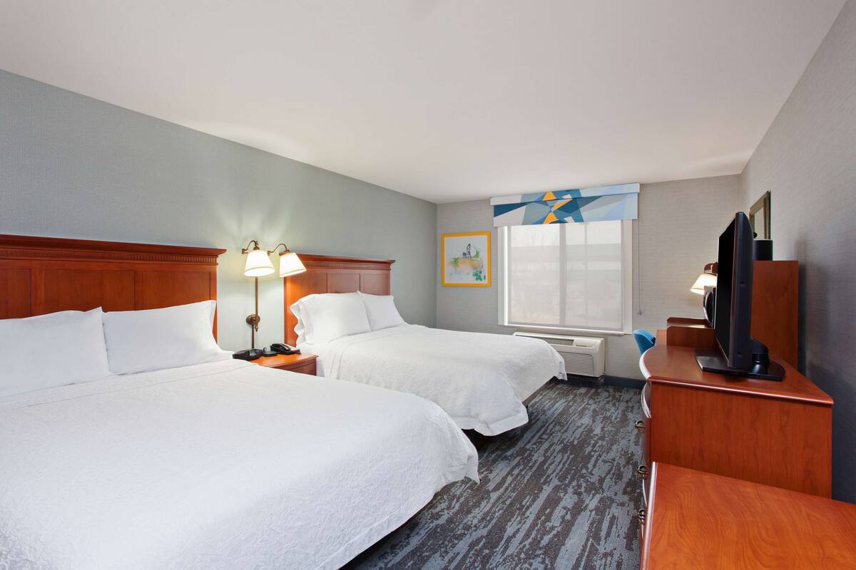 A hotel room with two beds, a desk with a TV, and a window with a geometric curtain.