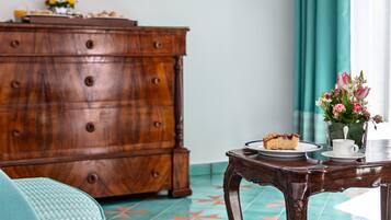 A wooden dresser, a table with a slice of cake, a cup, and a vase of flowers in a room with patterned floor tiles and teal curtains.