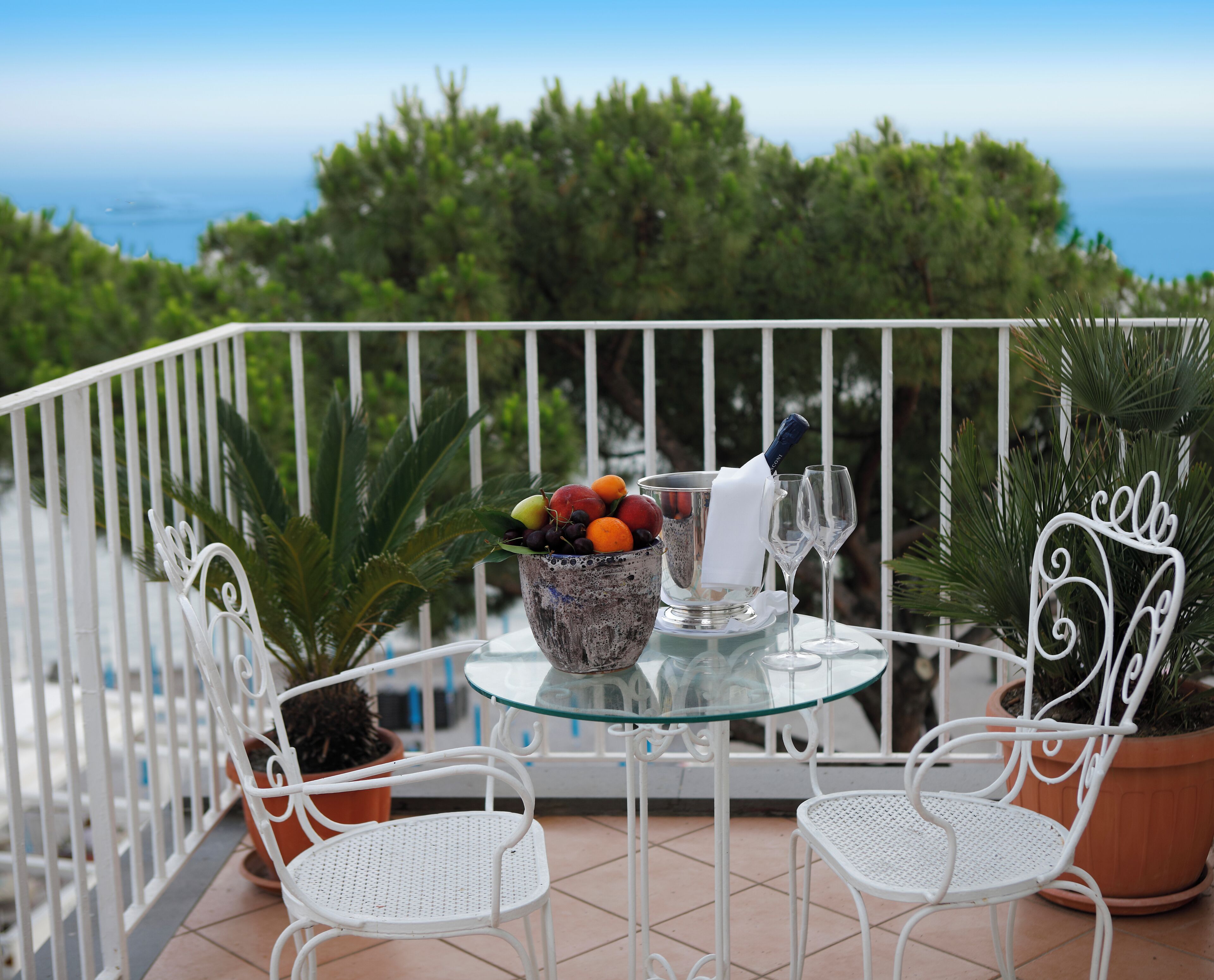 A balcony with a glass table, white metal chairs, a fruit bowl, a bottle of champagne, and potted plants.