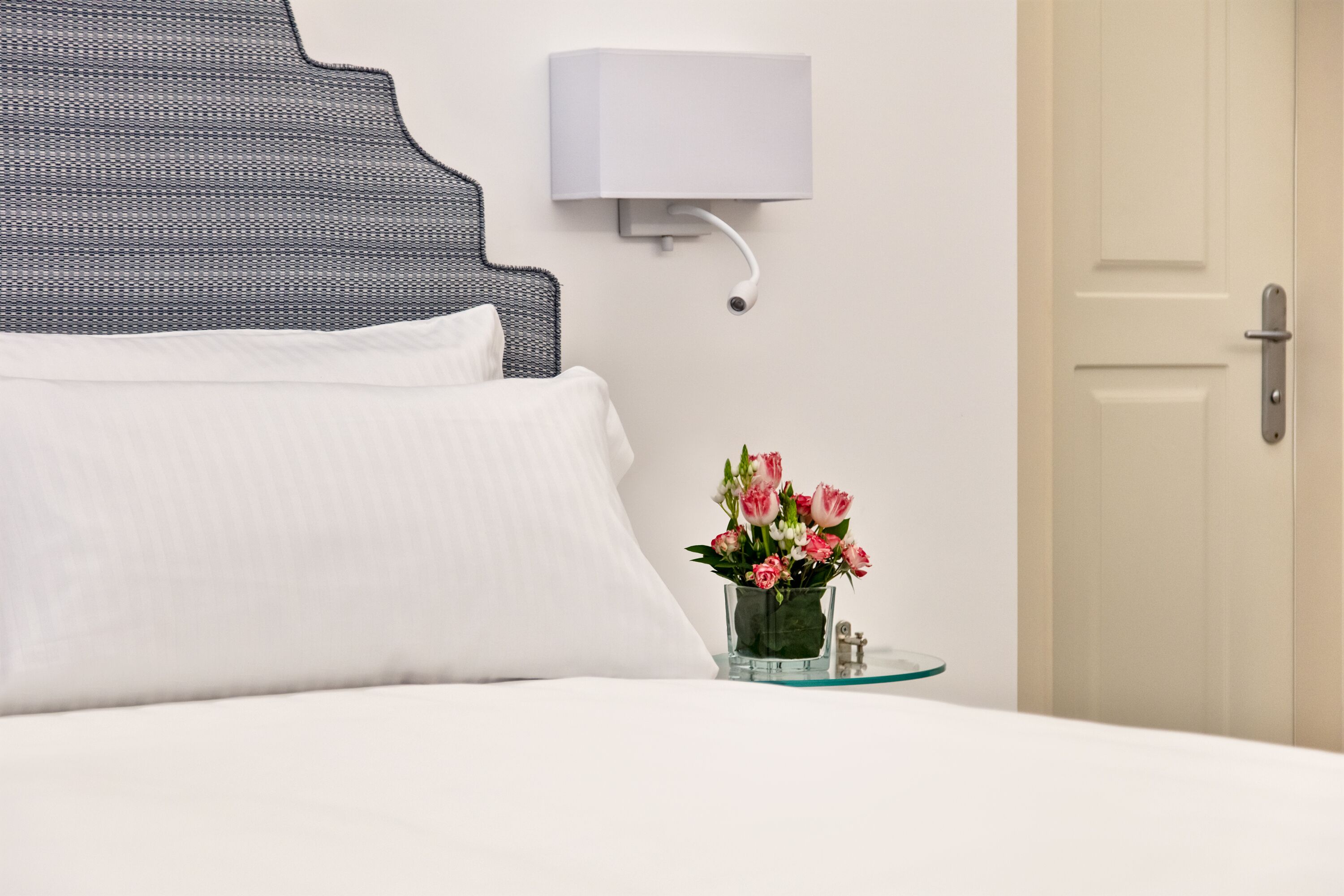 A neatly made bed with white linens, a wall-mounted light fixture, and a small glass vase with flowers on a bedside table.