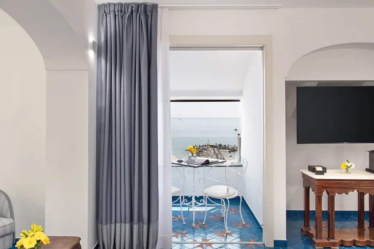 A room with a balcony, a dining table with chairs, a TV, and a small table with a vase of flowers.