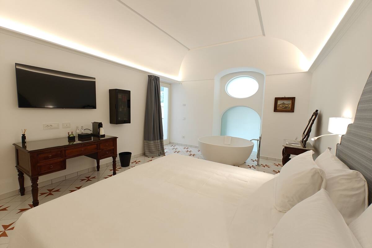 A modern hotel room with a large bed, a flat-screen TV, a vanity with a mirror, and a bathtub.