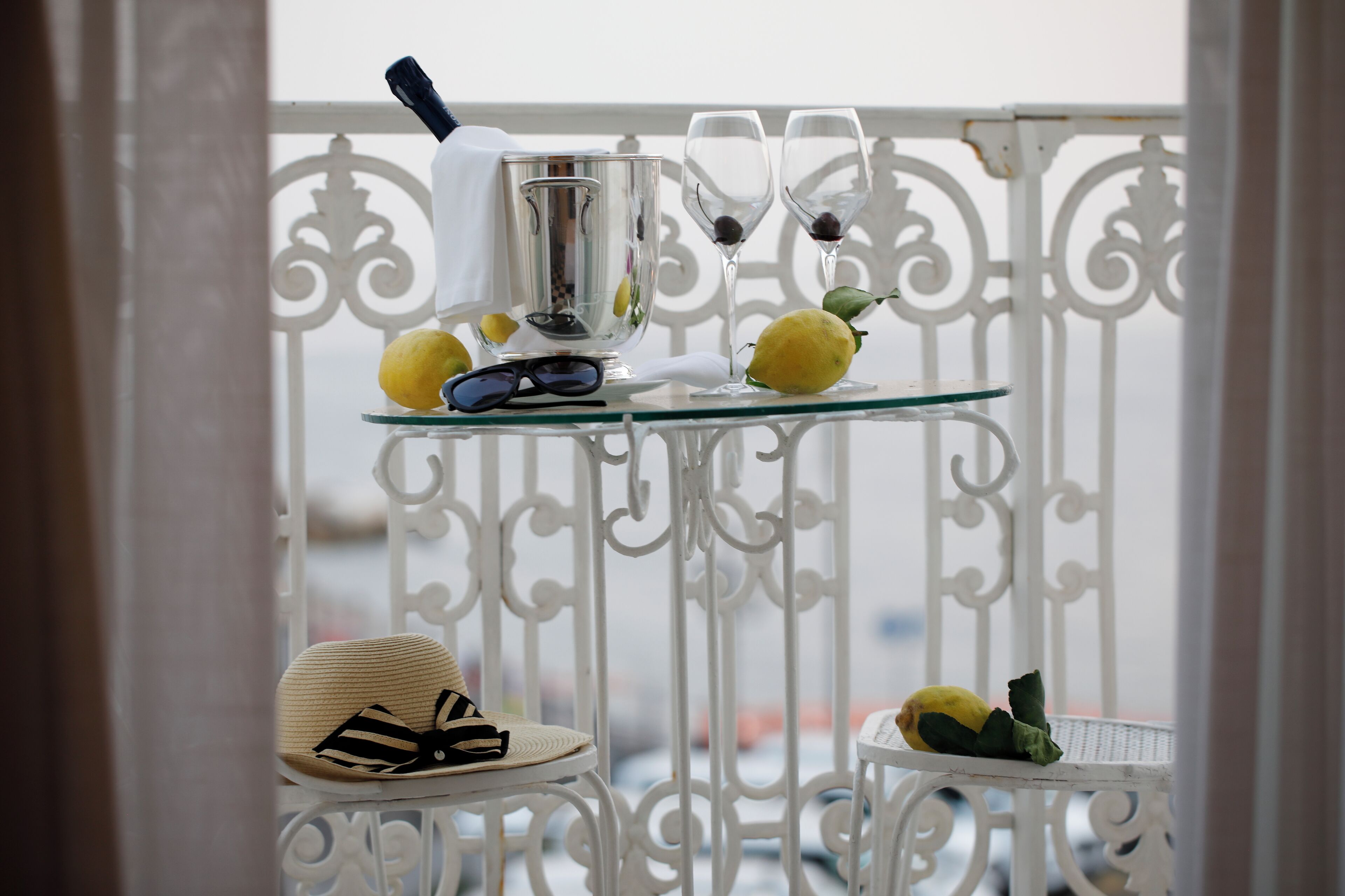 Een glazen tafel met een metalen karaf, twee wijnglazen en citroenen op een balkon met witte metalen hekken.