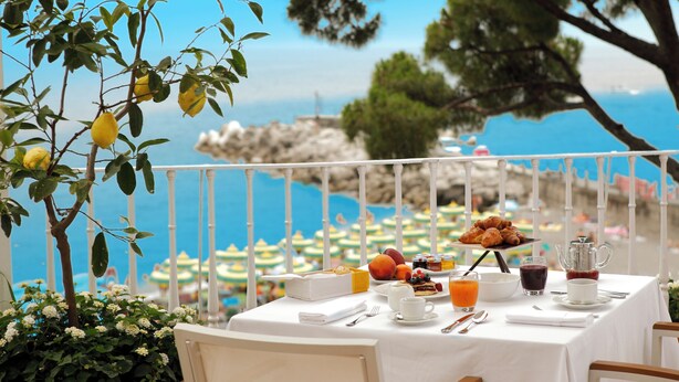 A table set with breakfast items, including croissants, fruit, and beverages, overlooking a scenic coastal view with umbrellas and a rocky shoreline. - Hotel Residence - Amalfi (Amalfi)