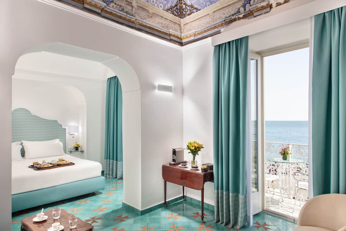 A hotel room with a bed, a desk with a vase of flowers, a view of the sea, and a balcony with chairs.