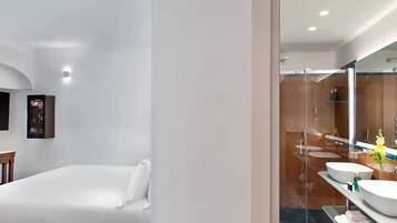 A modern hotel room with a bed, a large mirror, and a bathroom with a glass shower enclosure and two sinks.