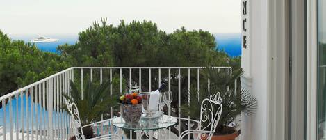 A balcony with a white railing, a table with chairs, potted plants, and a view of the sea.