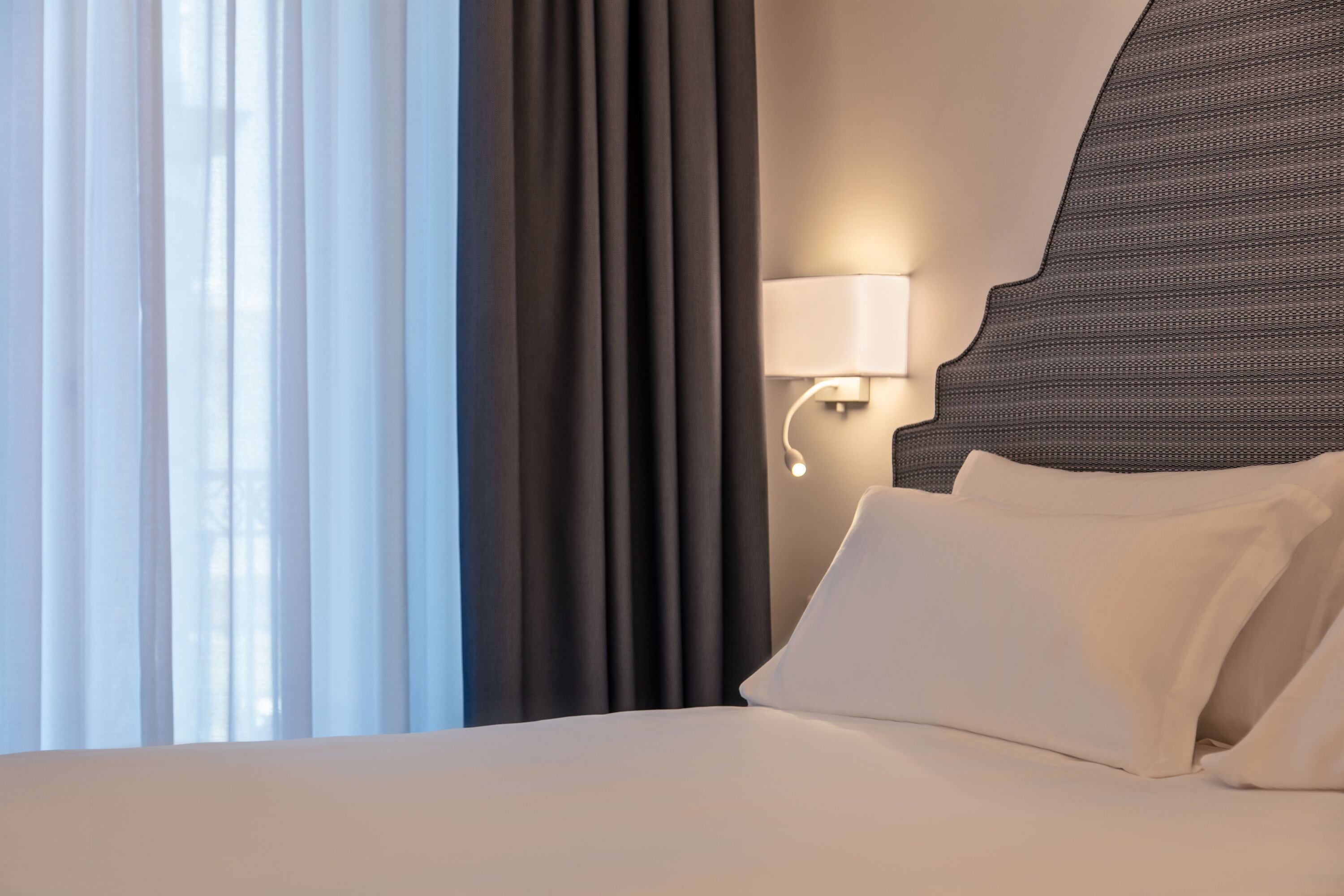 Habitación de hotel con cama, cortinas, lámpara de pared y ventana con cortinas translúcidas.