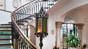 A grand staircase with ornate metal railings, a wooden table with chairs, and a potted plant. - Hotel Residence - Amalfi (Amalfi)
