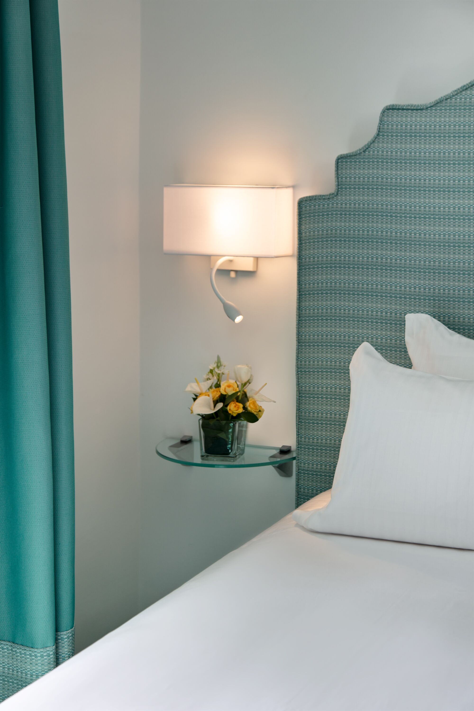 A neatly made bed with white linens, a bedside lamp, and a small glass table with a vase of flowers.
