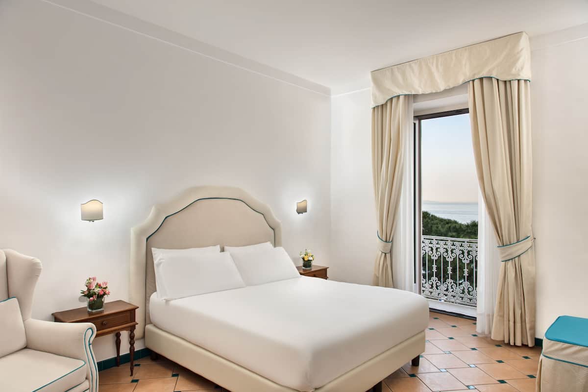 A hotel room with a large bed, a nightstand with a lamp, a chair, and a view of the sea through a balcony.