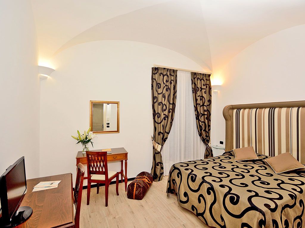 A hotel room with a bed, a desk, a chair, a small table, a mirror, and curtains.