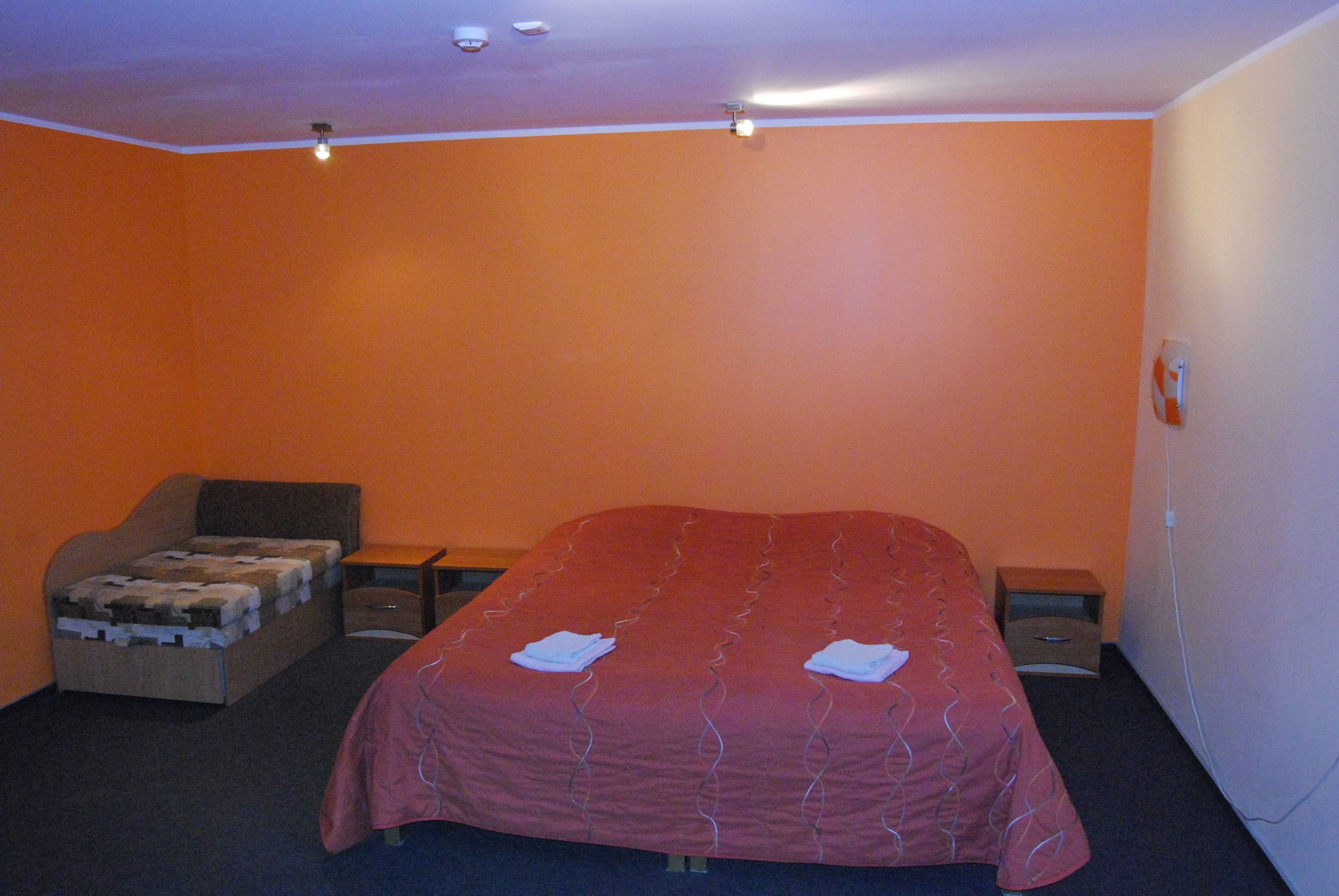 A bedroom with an orange wall, a bed with a red bedspread, a small sofa, and two bedside tables.