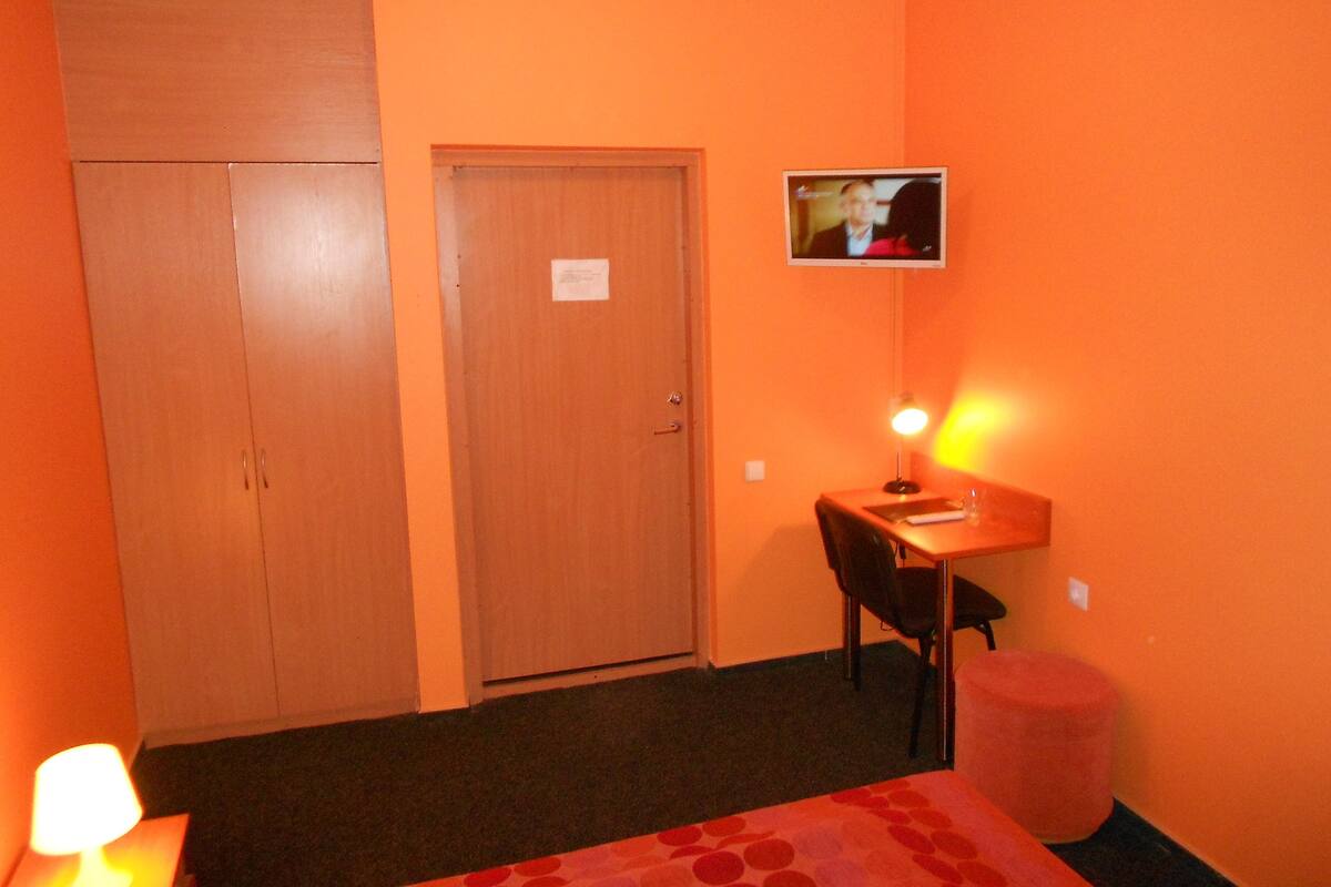 Une pièce comprenant une armoire en bois, un bureau avec une chaise, un téléviseur fixé au mur et un petit tabouret rouge.