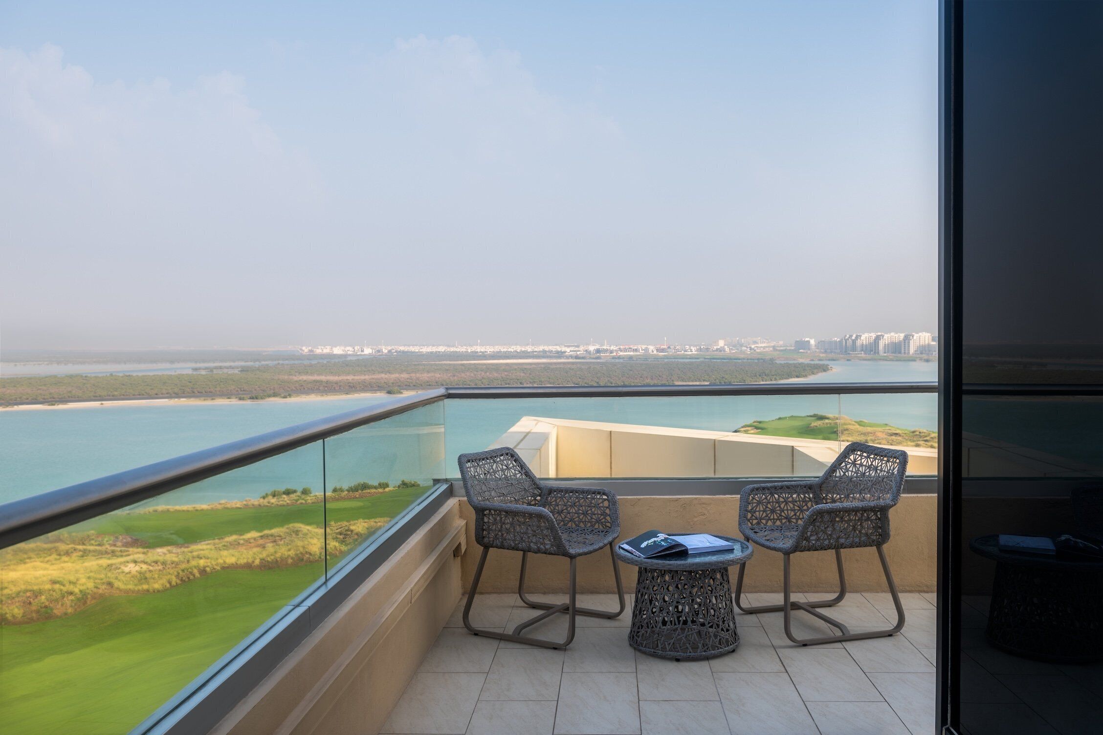 A balcony with two chairs and a small table, overlooking a coastal landscape with buildings in the distance.