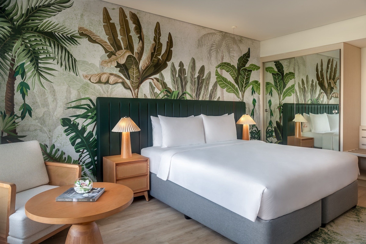 Una habitación de hotel con una cama grande, dos mesitas de noche con lámparas, una mesa redonda de madera y un mural en la pared con follaje tropical.