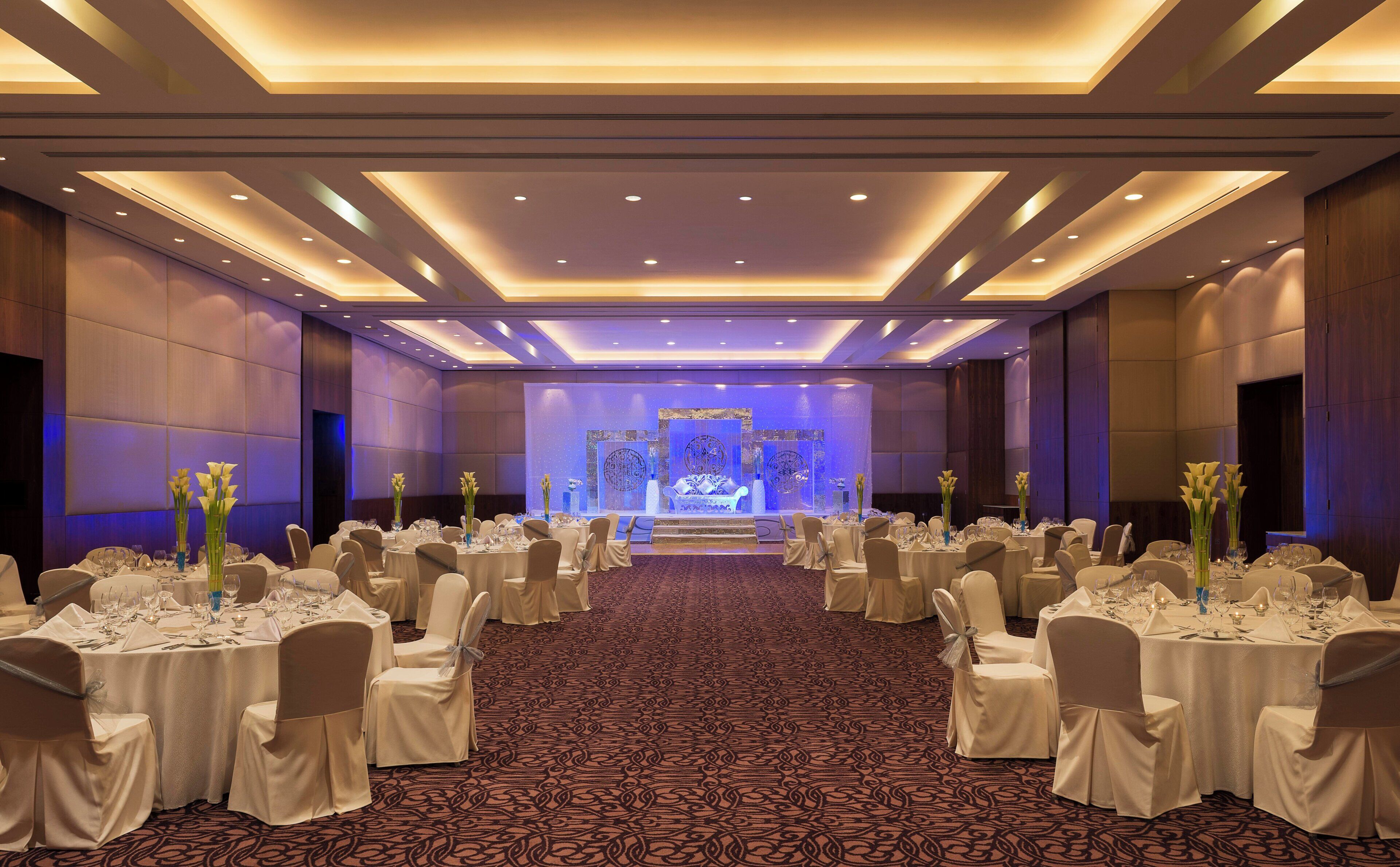 A banquet hall with round tables, white tablecloths, and chairs, a patterned carpet, and a stage with a blue-lit backdrop.