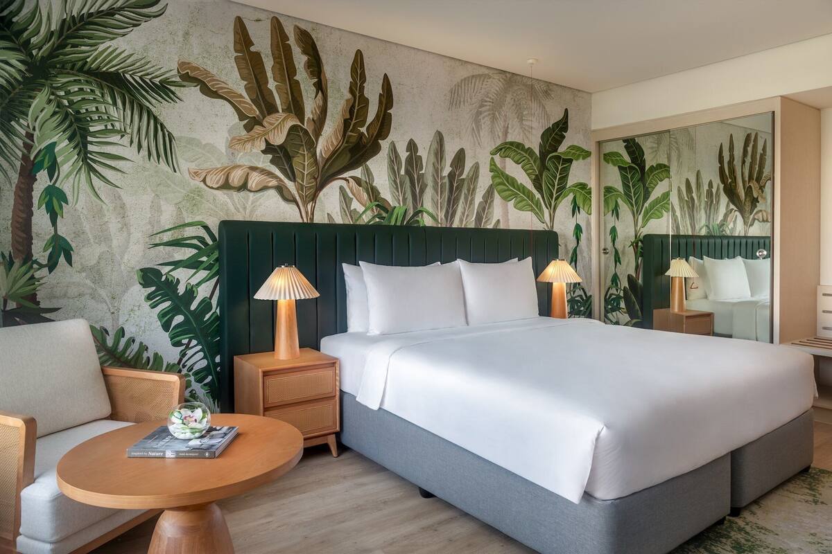 Una habitación de hotel con una cama grande, dos mesitas de noche con lámparas, una mesa redonda de madera y un mural en la pared con follaje tropical.