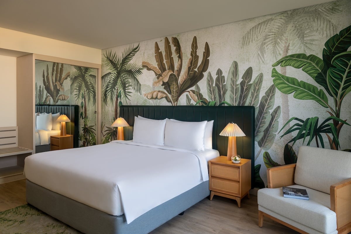 Habitación de hotel con una cama grande, dos mesitas de noche con lámparas, una silla y un mural de plantas tropicales.