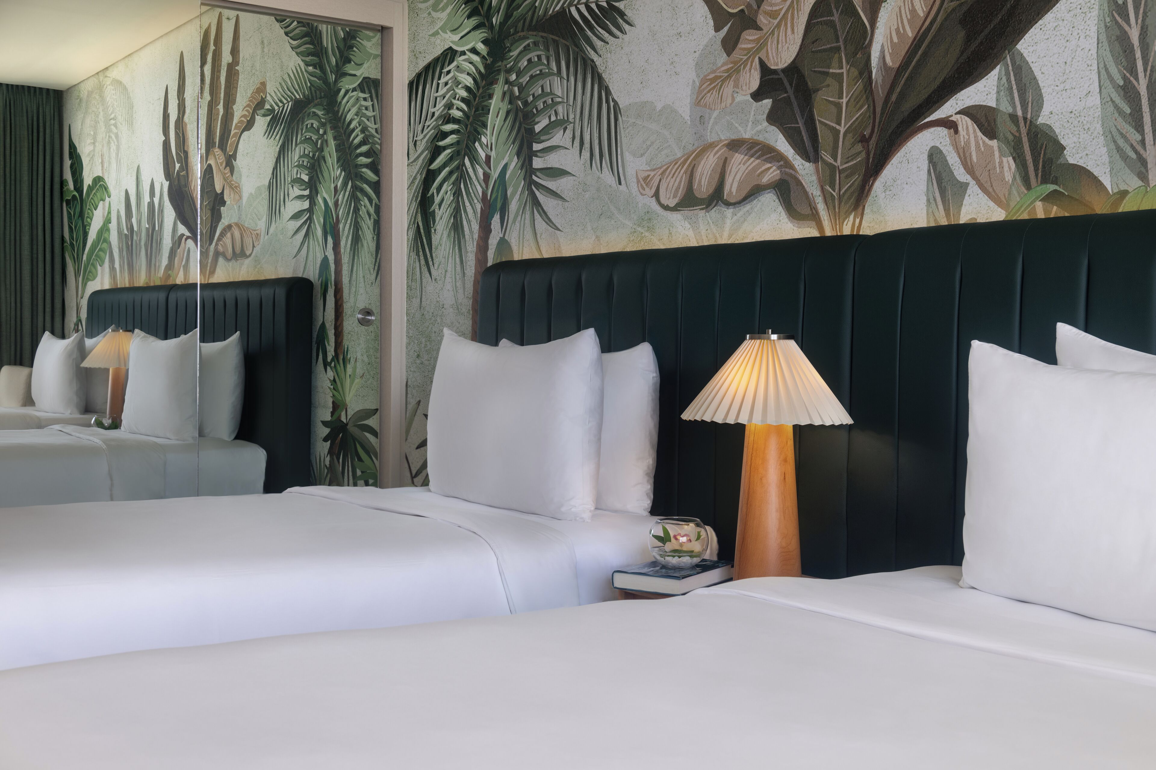 A hotel room with two beds, a large mural of tropical foliage on the wall, and a nightstand with a lamp.