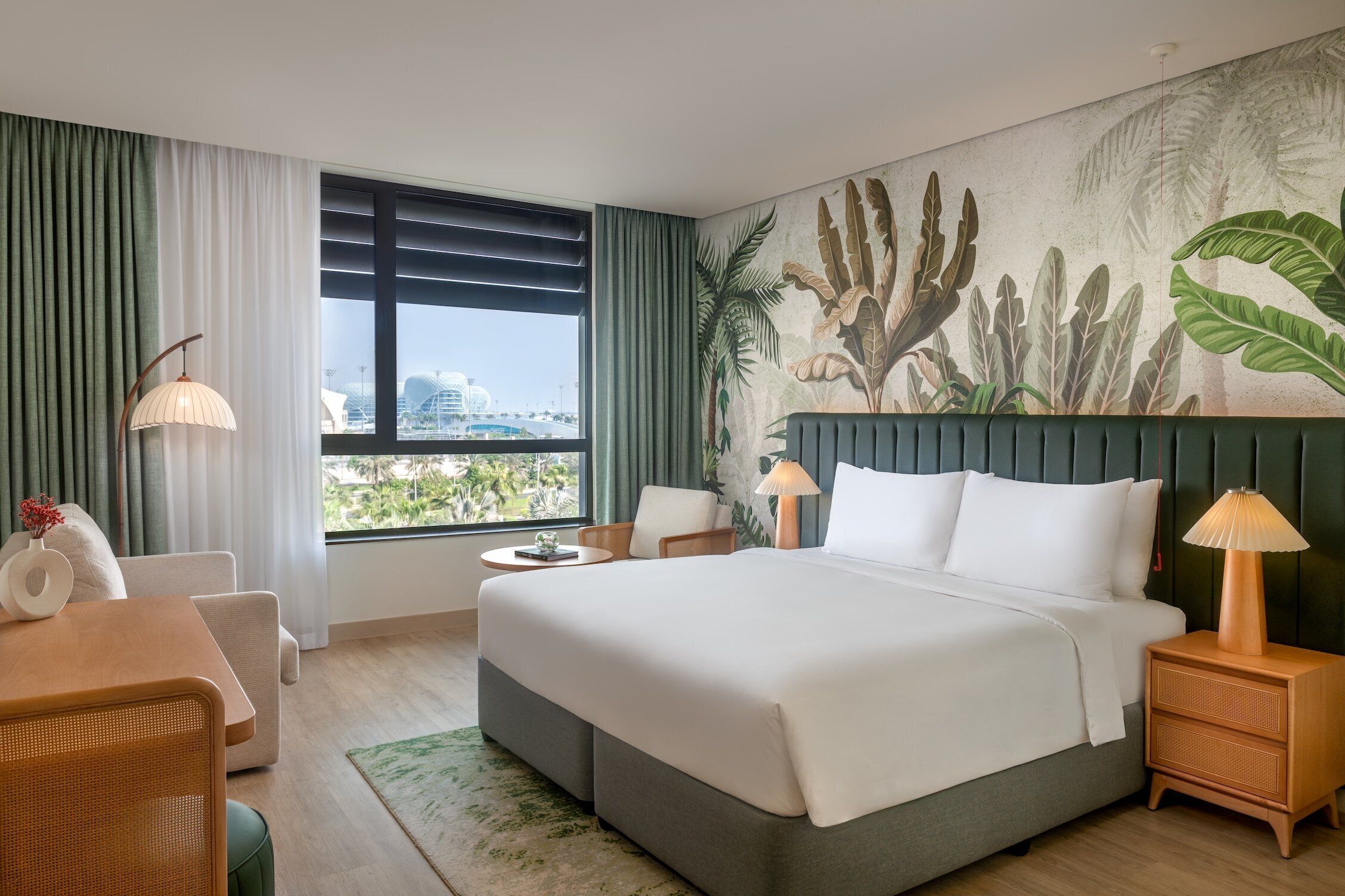Habitación de hotel con una cama grande, una mesa de comedor, una silla, una mesita auxiliar, una lámpara y vistas a la ciudad.