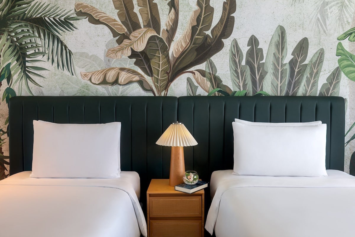 Una habitación con dos camas, una mesita de noche de madera con lámpara y una pared con un diseño de hojas.
