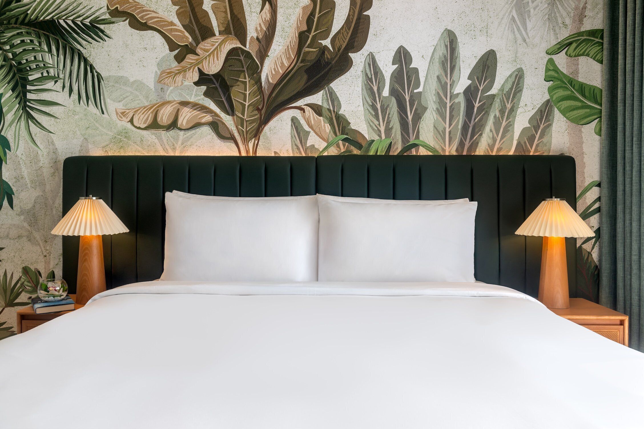 A bed with a dark headboard, two bedside lamps, and a tropical leaf mural on the wall.