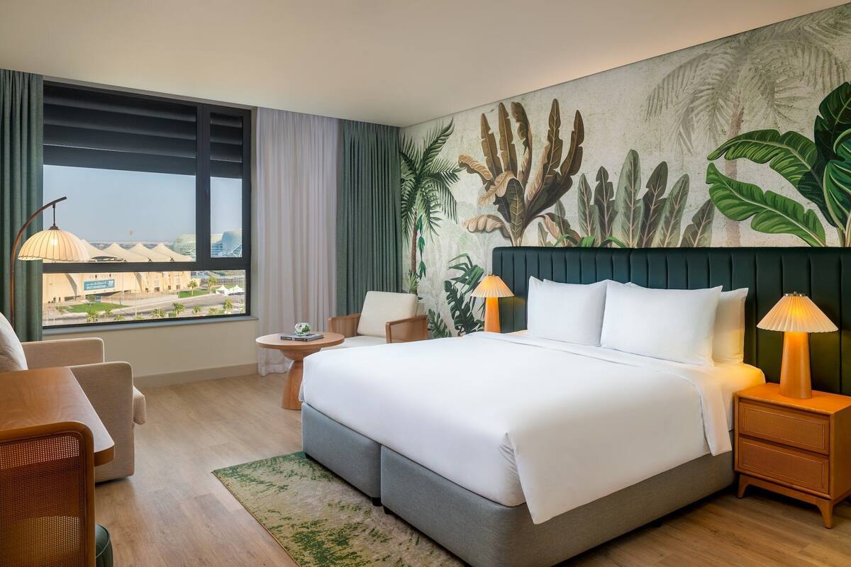 Habitación de hotel con una cama grande, dos mesitas de noche, una mesita redonda pequeña, una silla y vistas a un paisaje urbano a través de un ventanal grande.