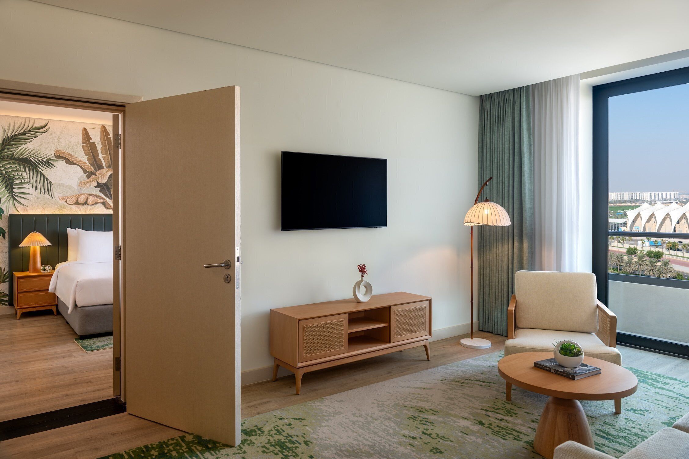 A hotel room with a bed, a TV, a seating area with a table and chair, a console table with a vase, and a large window with a view.