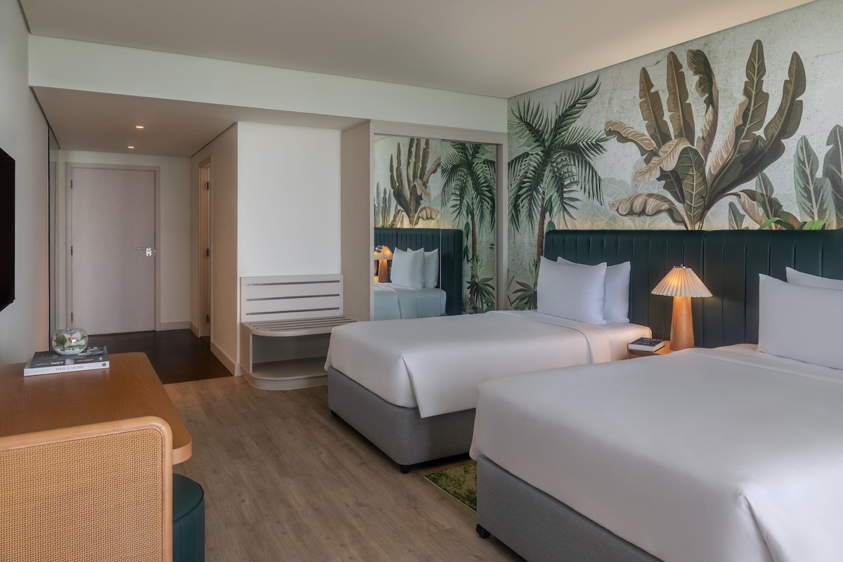 Habitación de hotel con dos camas, un escritorio, una silla, una lámpara y un mural de plantas tropicales.