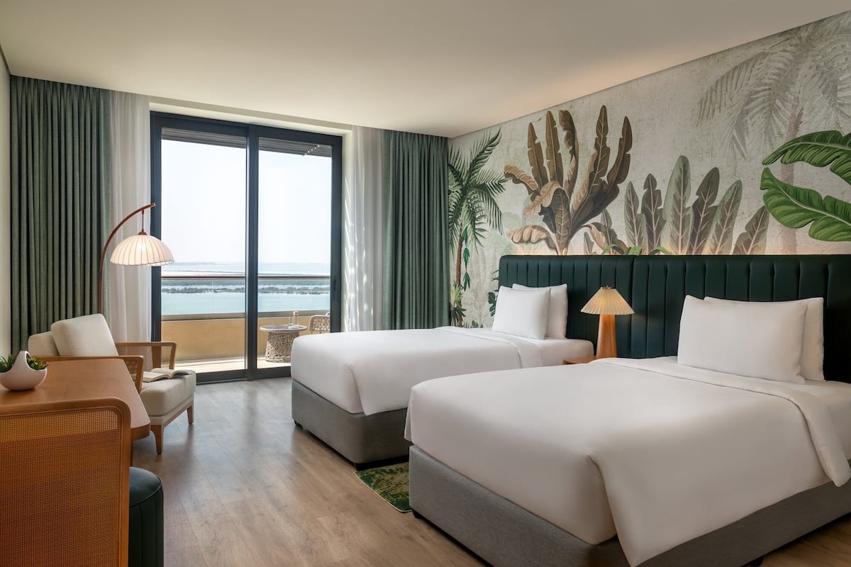 Habitación de hotel con dos camas, zona de comedor, ventana grande con vistas y un mural temático tropical.