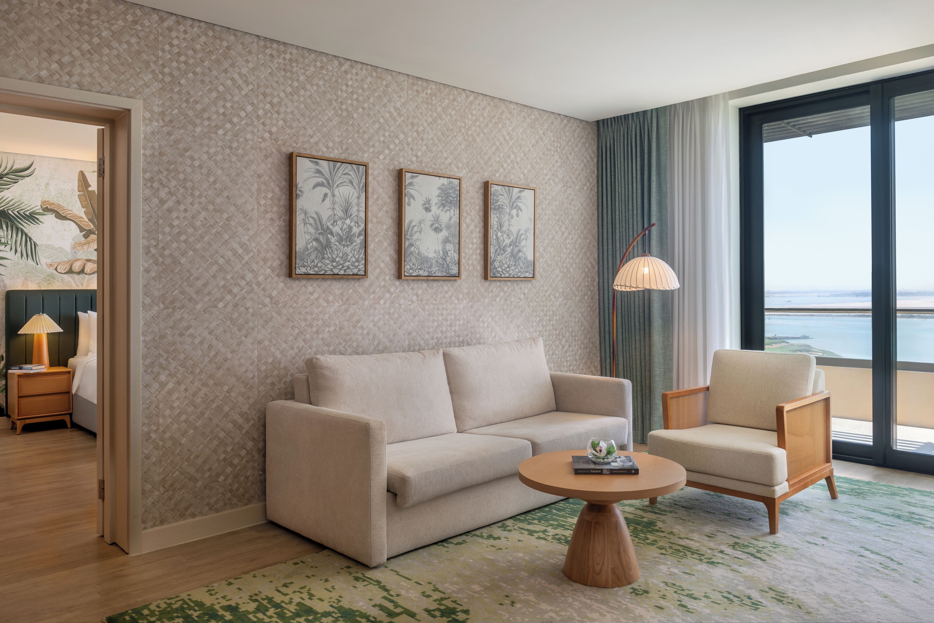 Una habitación de hotel moderna con un sofá color beige, una silla de madera, una mesa redonda de centro y un amplio ventanal con vistas al mar.