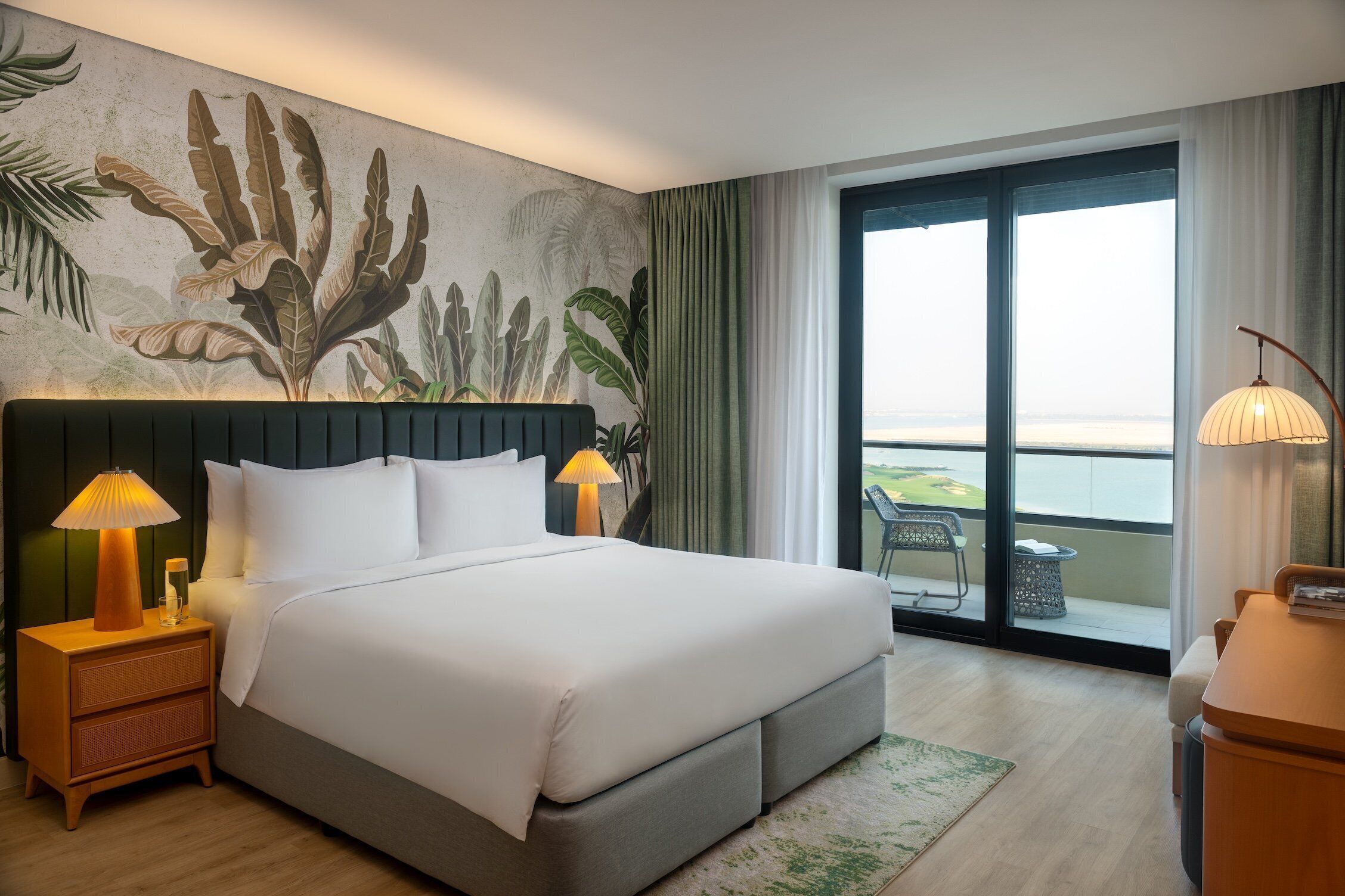 Ein Hotelzimmer mit Bett, zwei Nachttischlampen, einem Nachttisch, Blick ins Freie durch ein großes Fenster und einem Balkon mit einem Stuhl.