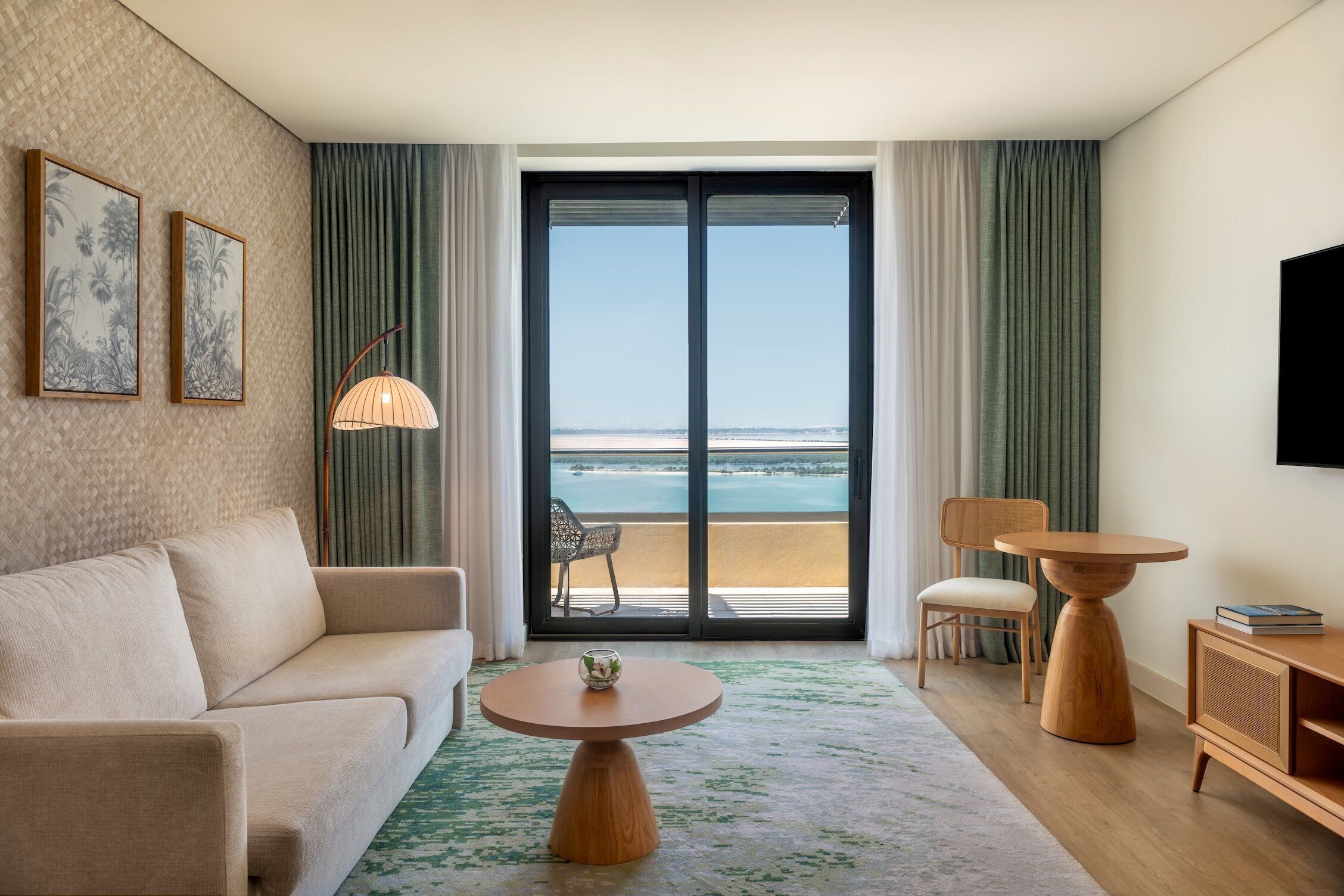 A modern hotel room with a sofa, two wooden side tables, a TV, and a balcony with a view of the ocean.