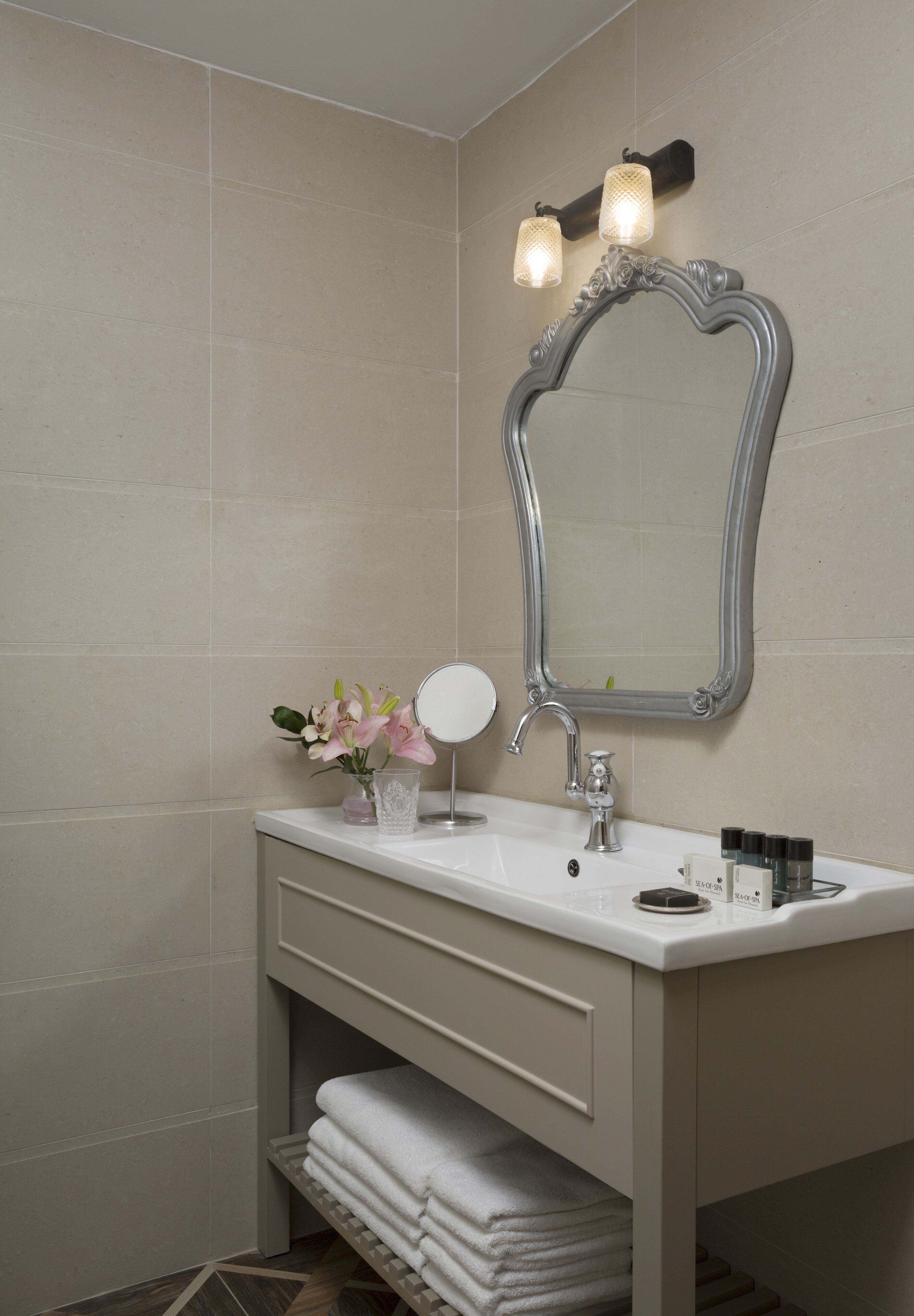 Een badkamer met een witte wastafel, een spiegel met een decoratief omlijsting, een handdoekrek en een kleine vaas met bloemen.