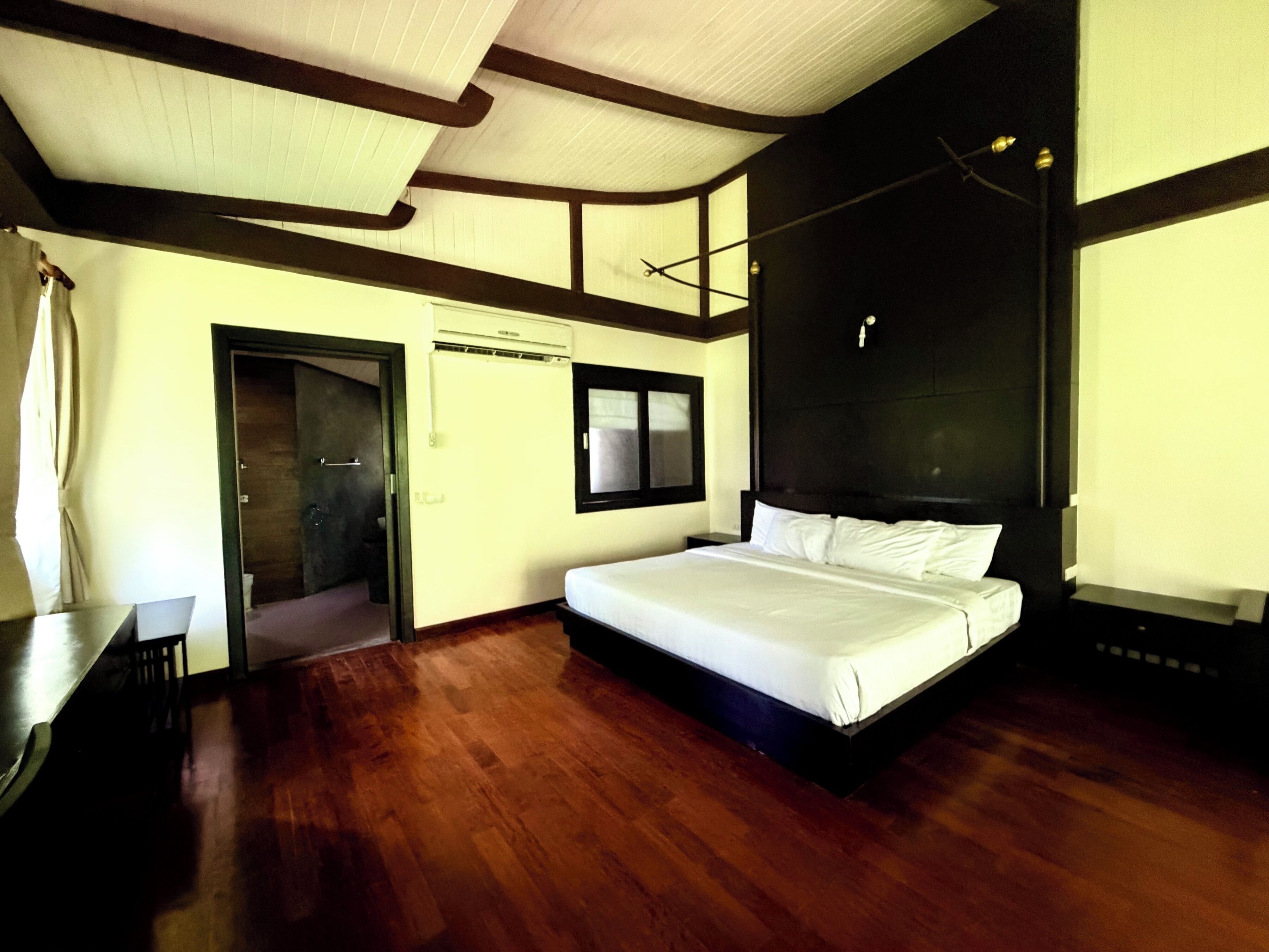 Un dormitorio con una cama grande, piso de madera, una puerta que da a otra habitación y una ventana.