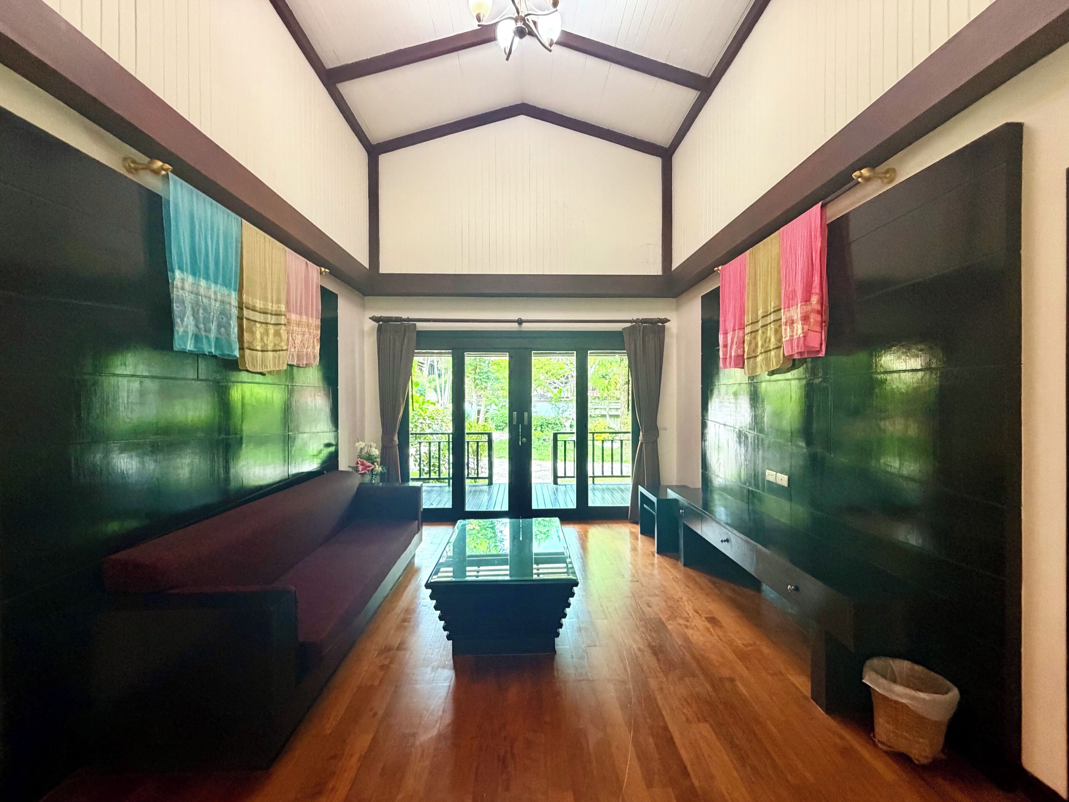 A spacious room with a high ceiling, wooden flooring, a central seating area, and large glass doors leading to an outdoor balcony.