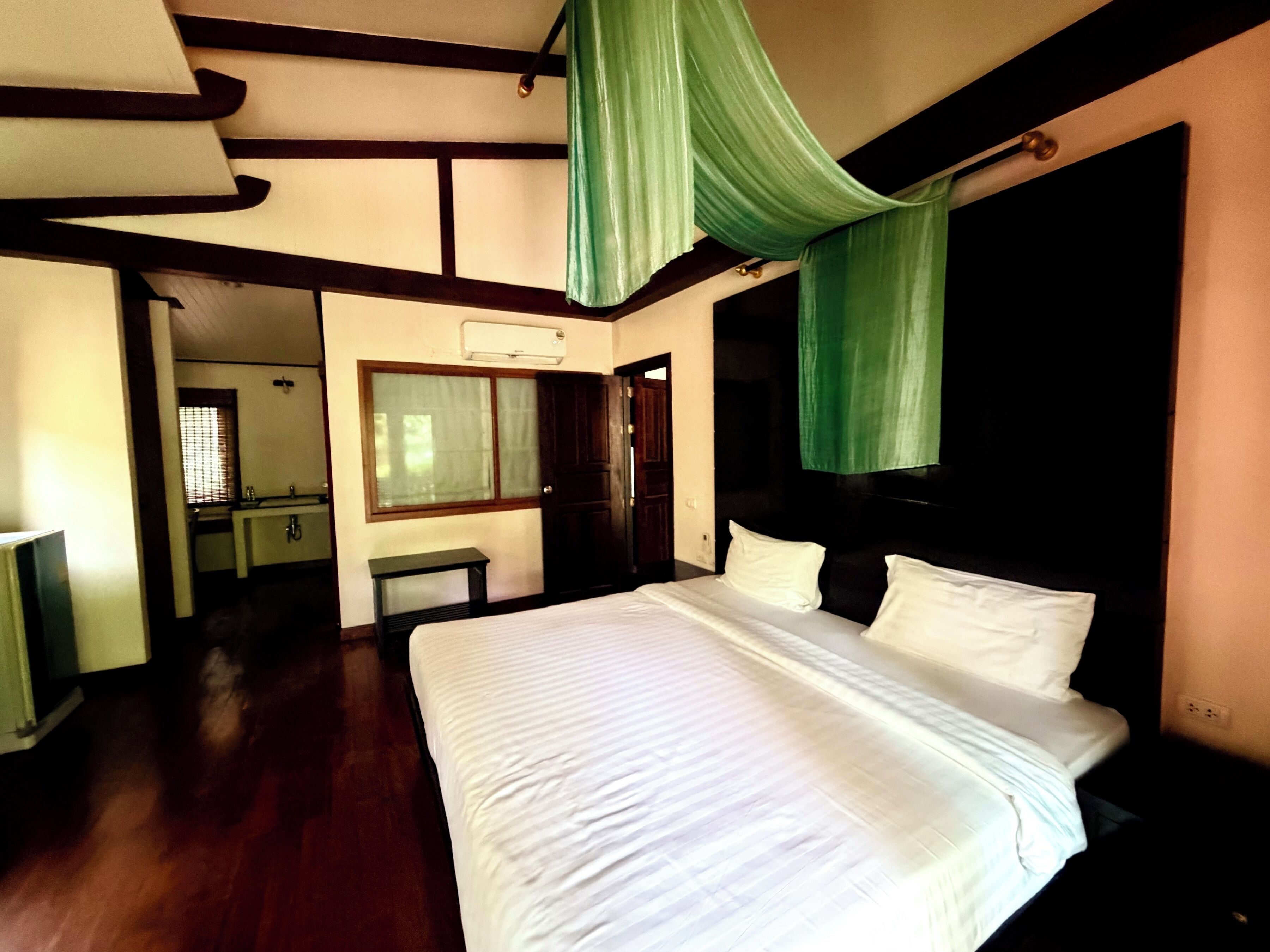A bedroom with a large bed, a wooden floor, a window with curtains, a bedside table, and a mirror.