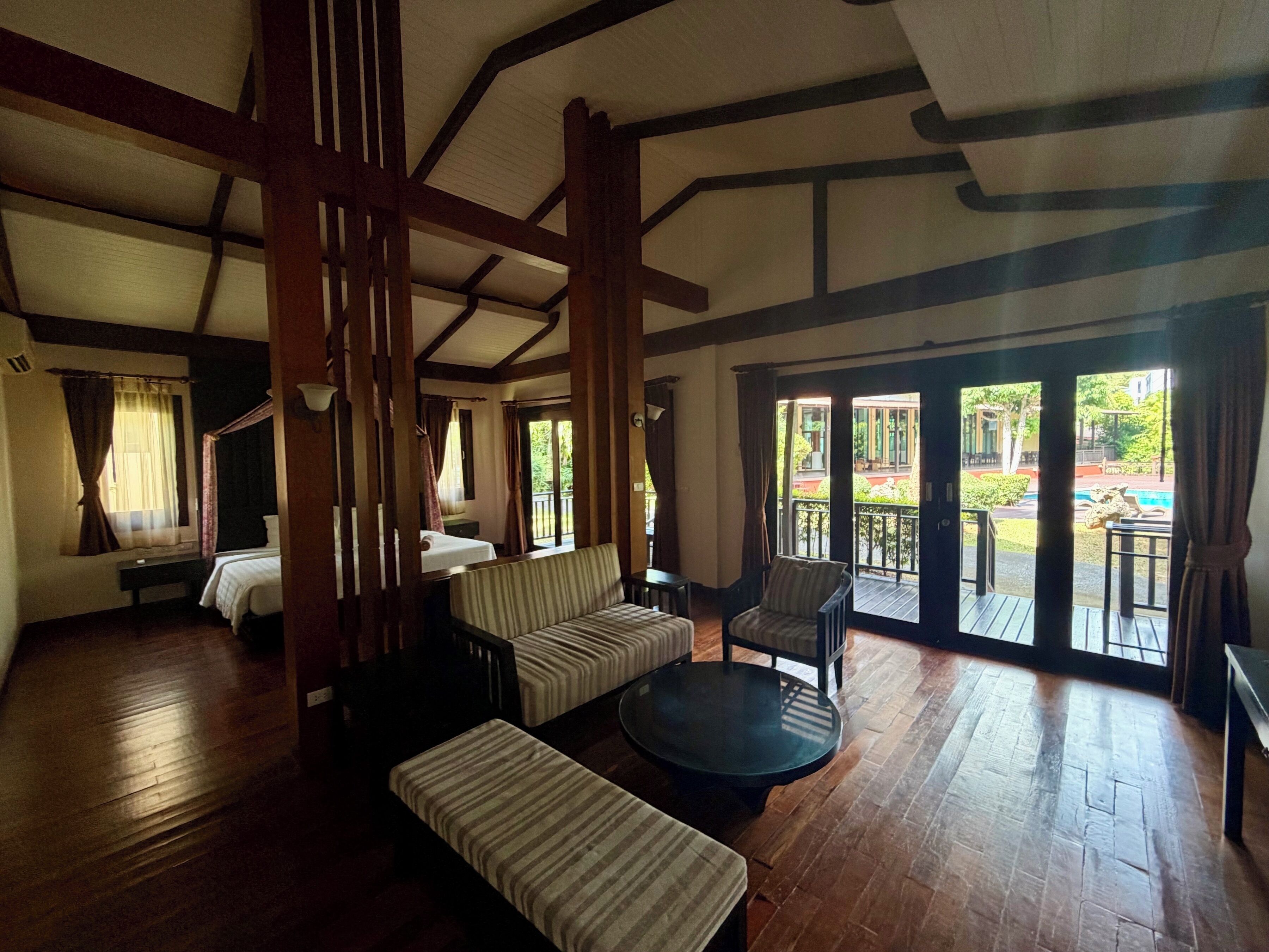 Una habitación con piso de madera, un sofá, una mesa redonda y una cama. Hay una ventana con cortinas y vista al exterior.