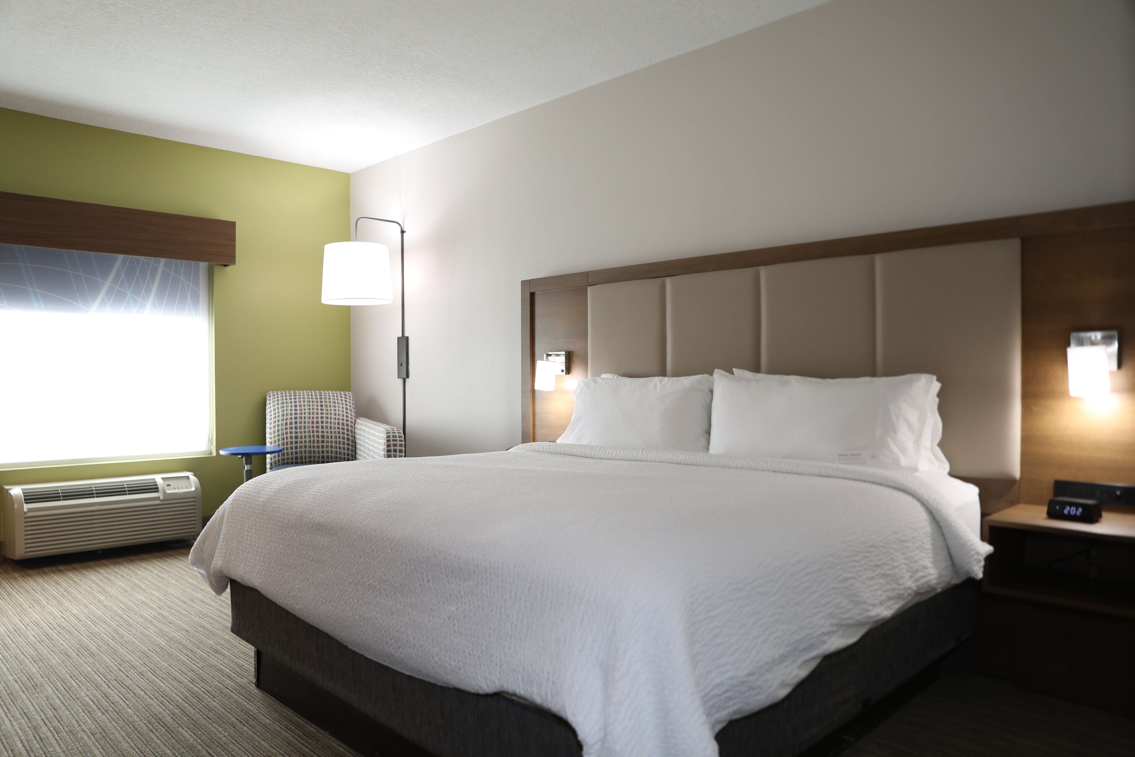 A hotel room with a large bed, a bedside table with a lamp, a chair, and a window with curtains.
